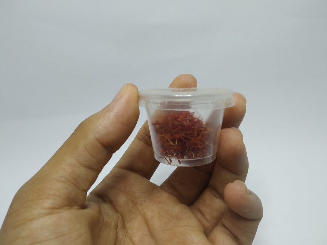 A hand holding a small clear lidded plastic cup filled with loose deep-red saffron threads with lighter yellow ends, against a plain white background