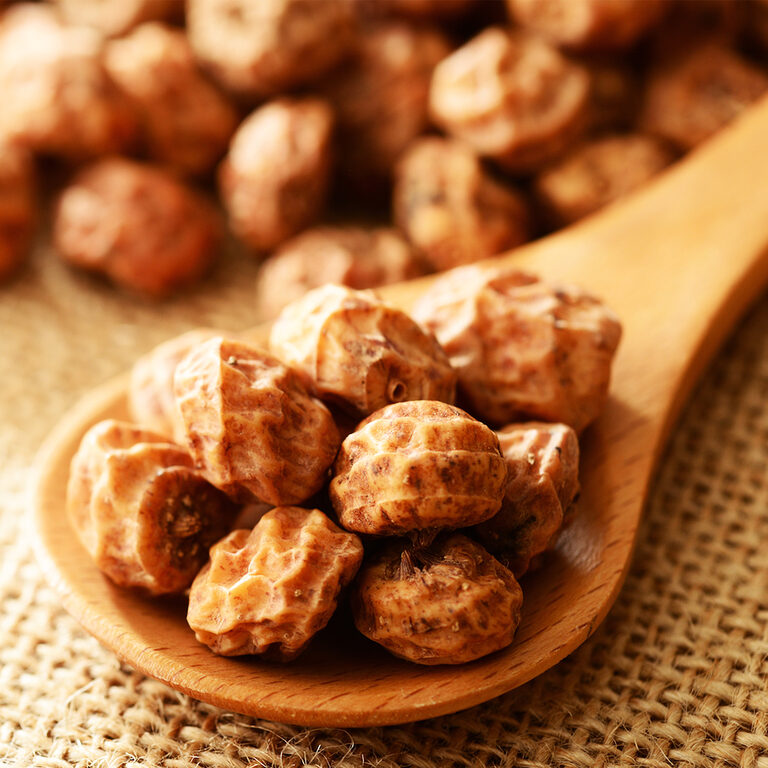 Close-up of wrinkled golden-brown dried tiger nuts heaped on a wooden spoon over burlap, with more tiger nuts blurred in the background