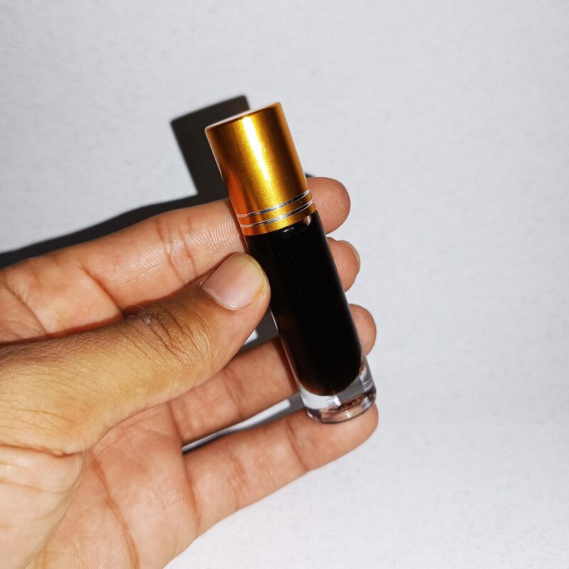 A hand holding a slim glass bottle of dark perfume oil with a gold cap against a pale background