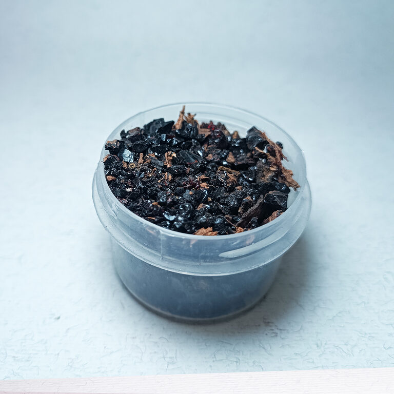 Clear cup filled with glossy black resin pieces and brown fibrous fragments on a pale surface.