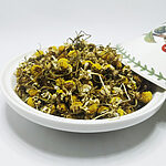 Dried chamomile flower heads with yellow centres, cream petals and thin stems heaped in a white ceramic dish, a floral-painted lid resting at the side