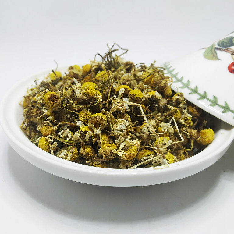 Dried chamomile flower heads with yellow centres, cream petals and thin stems heaped in a white ceramic dish, a floral-painted lid resting at the side
