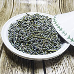 Dried grey-violet lavender buds with fine green stem pieces heaped in a white ceramic dish, its leaf-patterned lid resting beside it on a wooden surface
