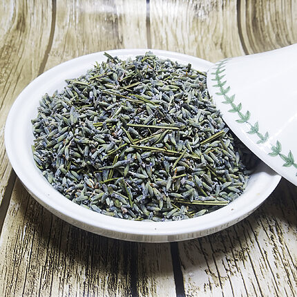 Dried grey-violet lavender buds with fine green stem pieces heaped in a white ceramic dish, its leaf-patterned lid resting beside it on a wooden surface