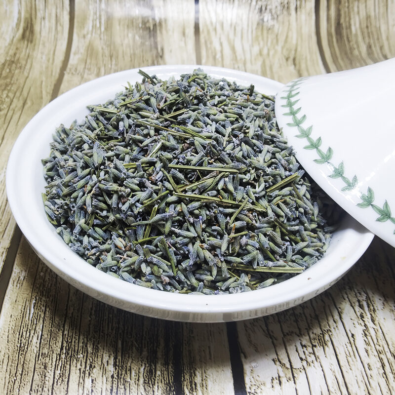 Dried grey-violet lavender buds with fine green stem pieces heaped in a white ceramic dish, its leaf-patterned lid resting beside it on a wooden surface