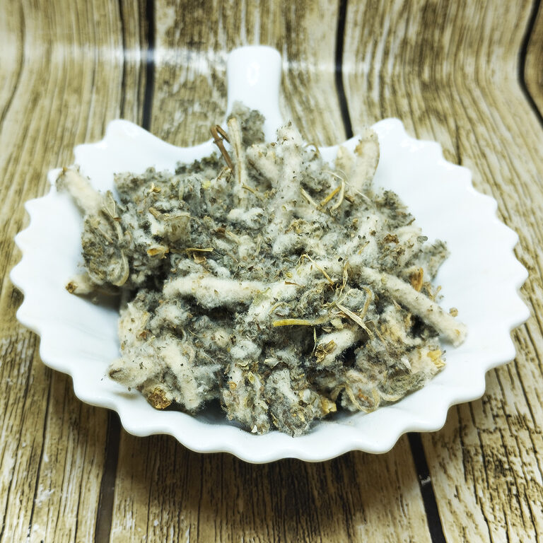 Dried white horehound with woolly silver-grey leaves and white downy stems heaped in a scalloped white ceramic dish on a wood-effect background
