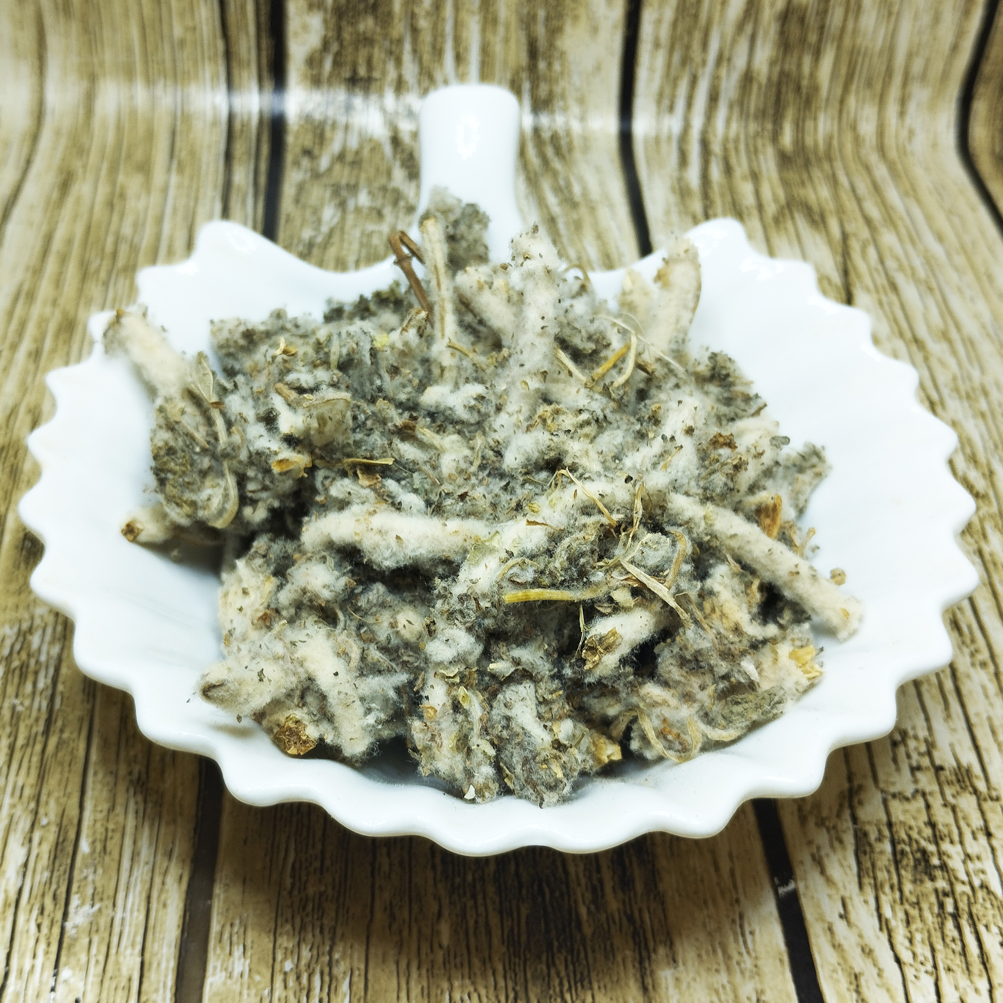 001-moroccan-marrubium-variation Dried white horehound with woolly silver-grey leaves and white downy stems heaped in a scalloped white ceramic dish on a wood-effect background