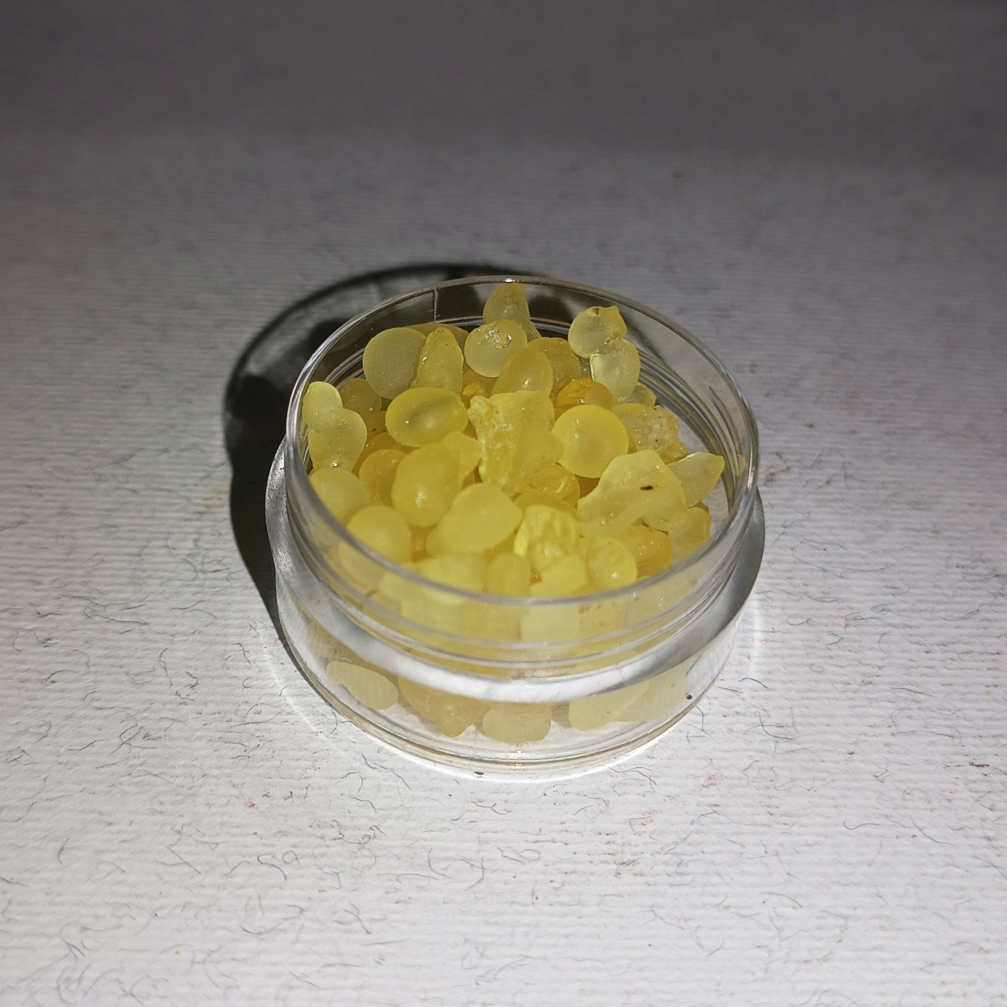 001-moroccan-natural-mastic-resin-whole-crystals-variation Pale yellow mastic resin tears, frosted and partly translucent, filling a small clear round jar on a white textured surface.