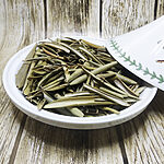 Whole dried olive leaves in sage-green and olive tones piled in a white ceramic tagine dish, its leaf-patterned lid tilted beside it on a weathered wooden surface