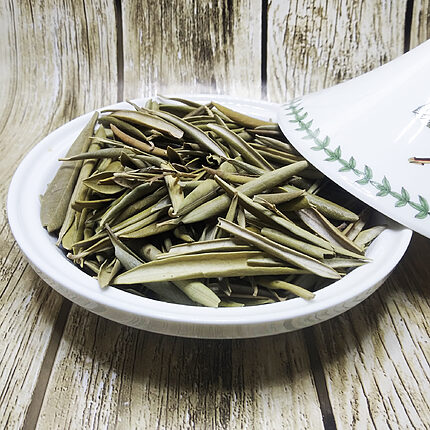 Feuilles d'olivier séchées entières, vert sauge et olive, empilées dans un plat à tajine en céramique blanche, son couvercle à motif de feuilles incliné à côté sur un fond en bois patiné