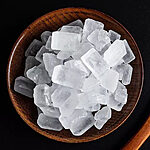 Top view of translucent white rock sugar crystal chunks piled in a round wooden bowl on a black background, with the edge of a wooden spoon at the right