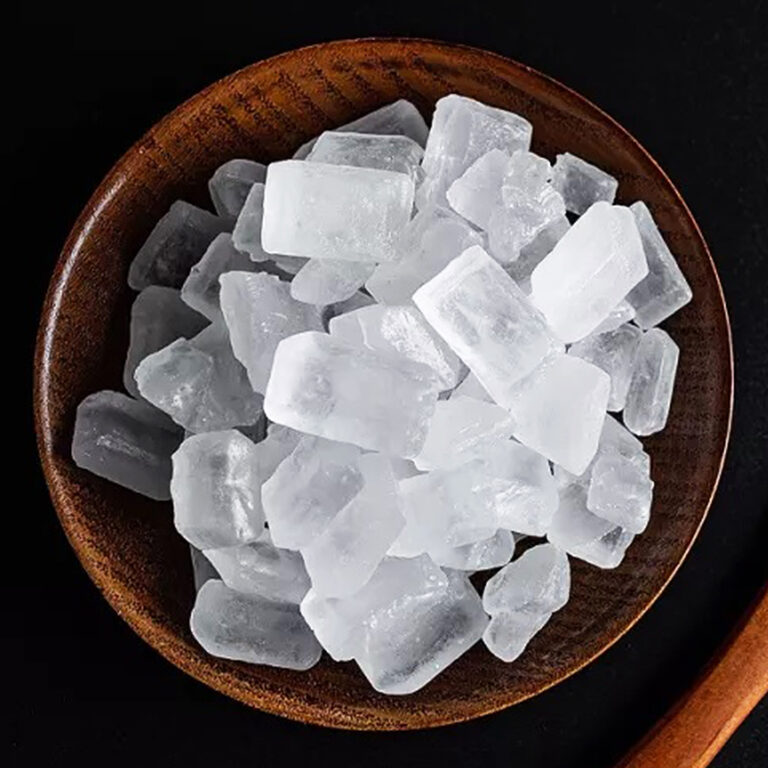 Top view of translucent white rock sugar crystal chunks piled in a round wooden bowl on a black background, with the edge of a wooden spoon at the right