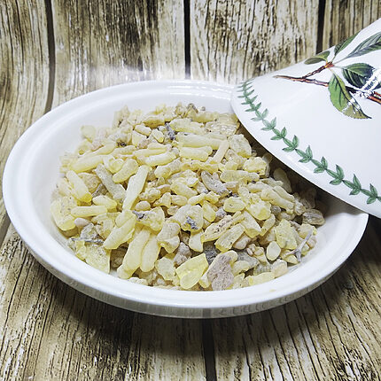 Larmes de résine allongées jaune pâle avec quelques fragments d'écorce dans un plat blanc, à côté d'un couvercle peint de feuilles vertes sur du bois patiné