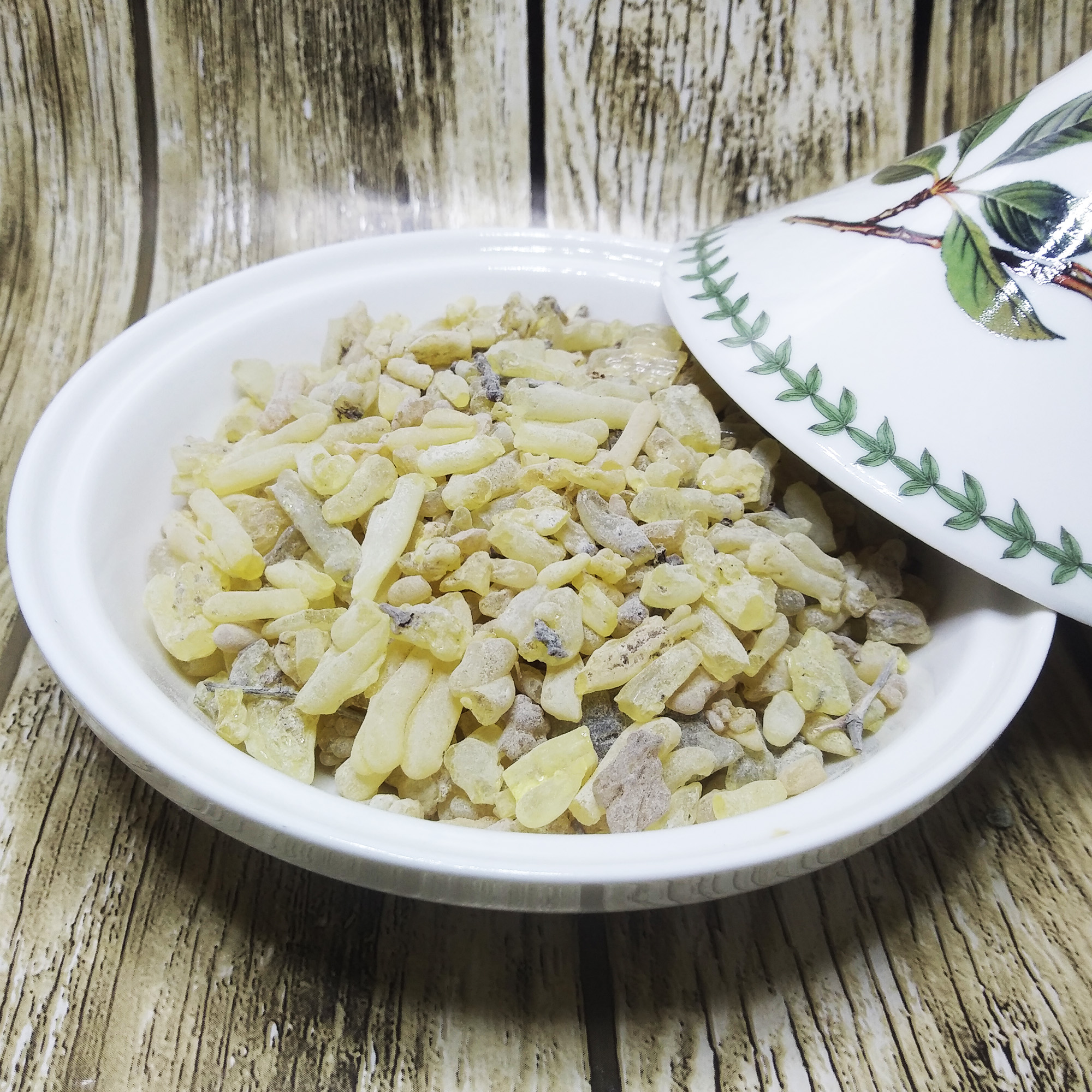 001-moroccan-sandarac-incense-variation Larmes de résine allongées jaune pâle avec quelques fragments d'écorce dans un plat blanc, à côté d'un couvercle peint de feuilles vertes sur du bois patiné