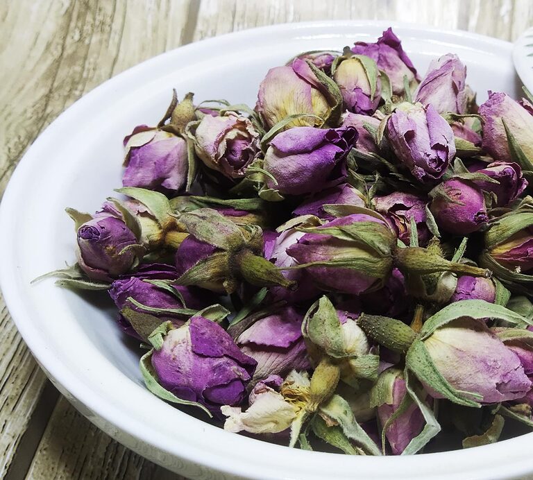 Whole purple-pink dried rose buds with green sepals in a white bowl on a wooden surface