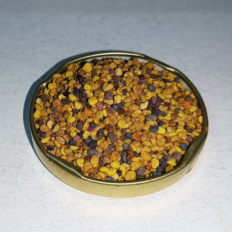 Mixed yellow, orange, brown and purple bee pollen granules in a shallow brass dish.