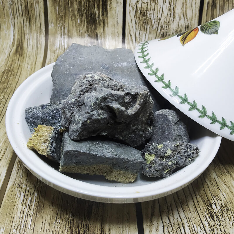 Irregular charcoal-black mineral chunks with exposed yellow areas on a white plate beside a white floral-patterned dish