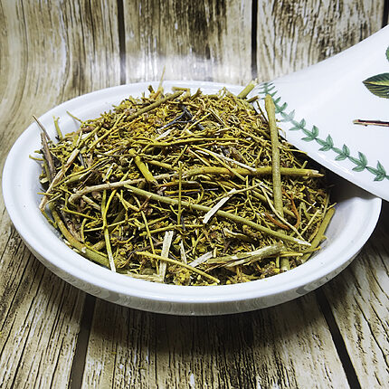 Bowl filled with green dried rue stems, leaves and flower tips beside a white spoon