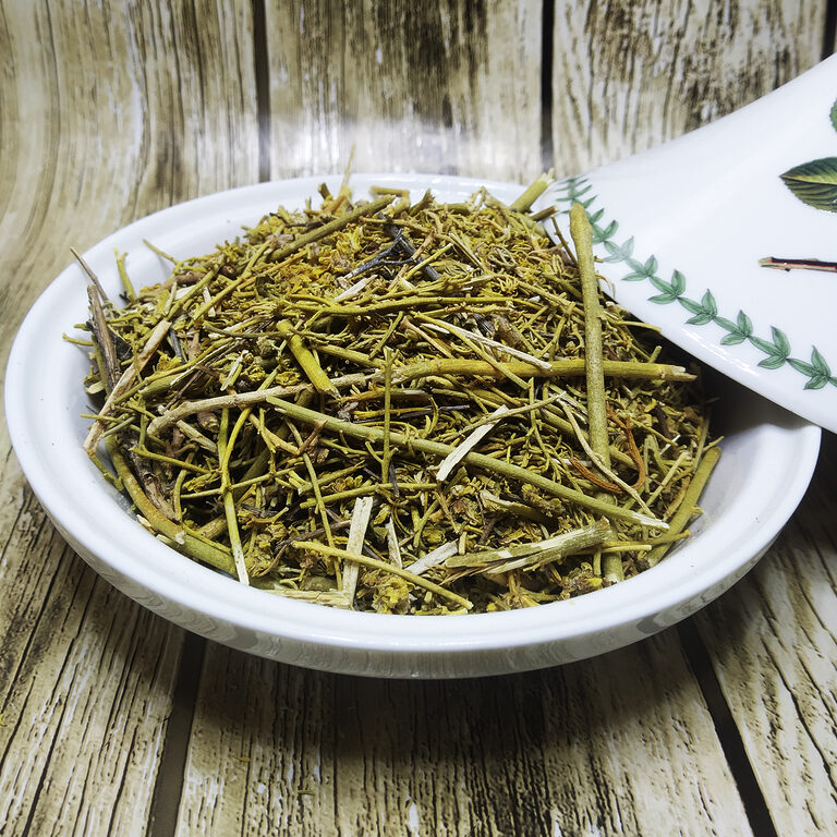 Bowl filled with green dried rue stems, leaves and flower tips beside a white spoon