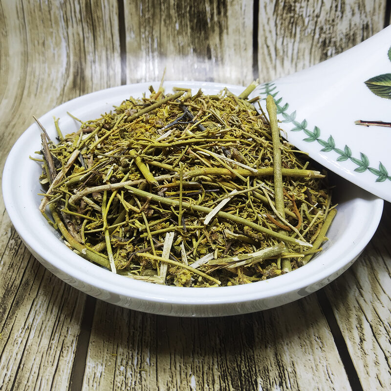 Bowl filled with green dried rue stems, leaves and flower tips beside a white spoon