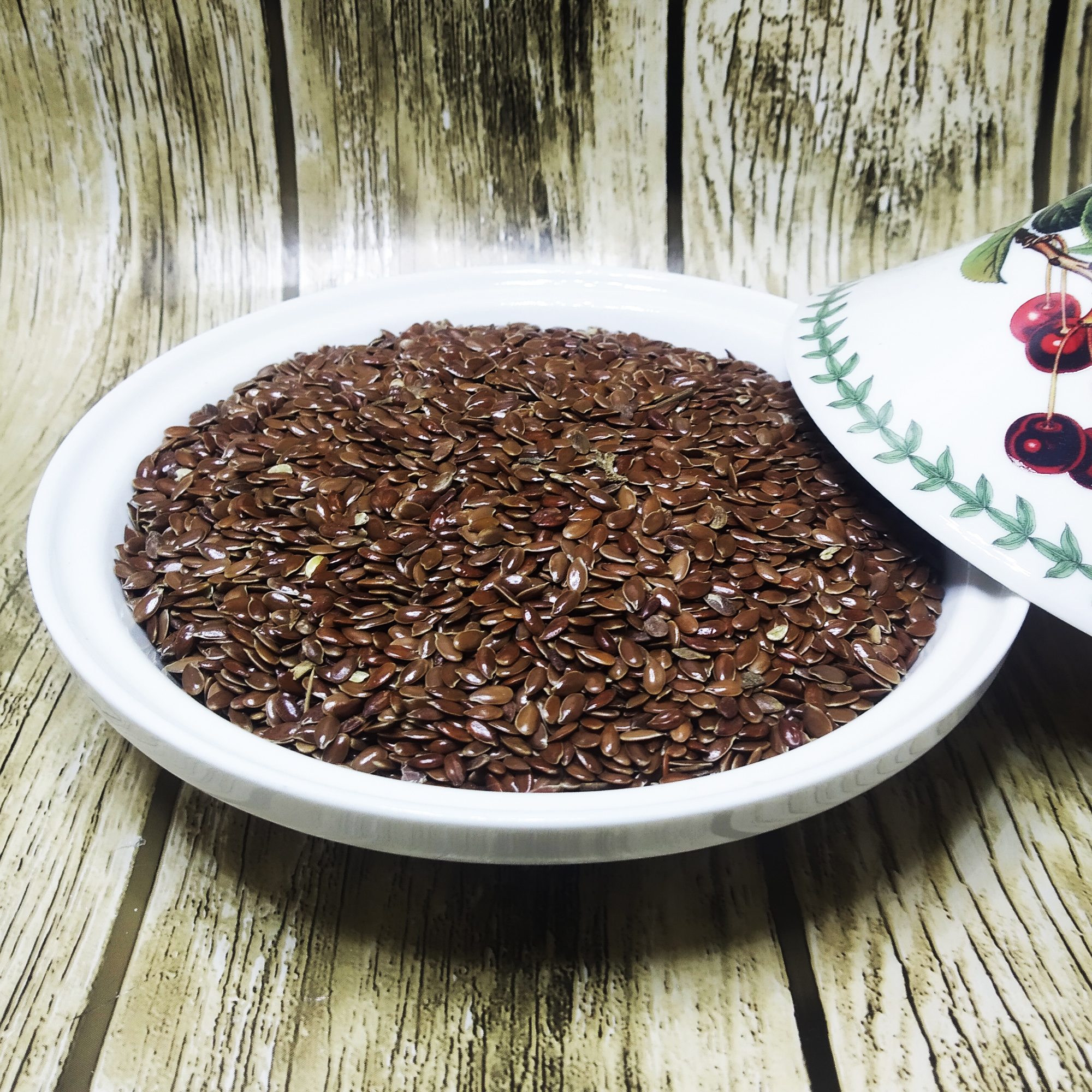 001-v-61 Glossy brown flax seeds piled in a white dish on a wood-grain surface