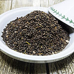 Close-up of loose dark Peganum harmala seeds piled in a white dish on a wood-grain surface.