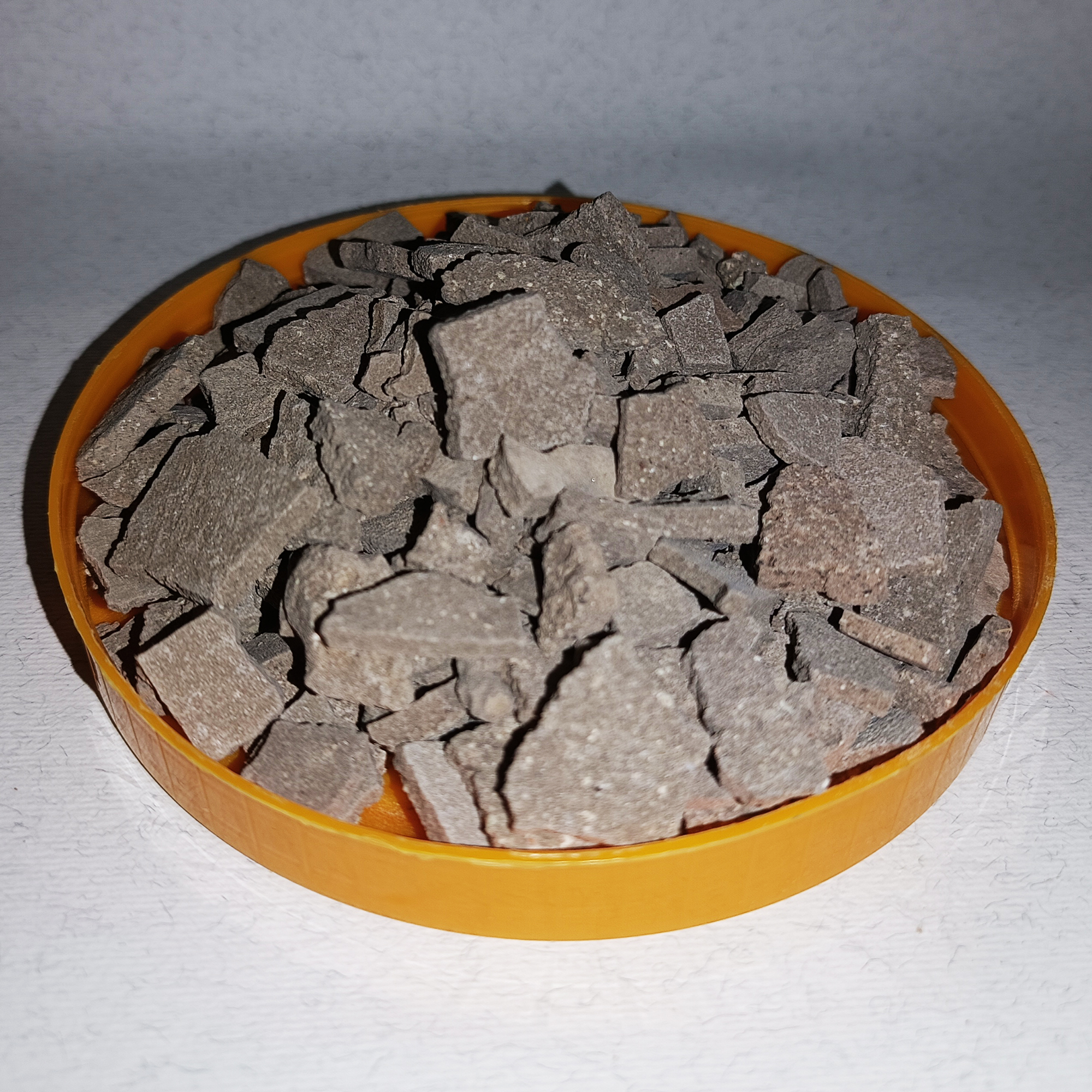 001-v-66 Grey-brown dry rhassoul clay pieces in a round orange dish.