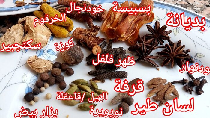 Whole spices laid out on a white floral plate with red Arabic labels, including ginger, turmeric, galangal, mace, star anise, nutmeg, long pepper, cardamom, cinnamon and cloves